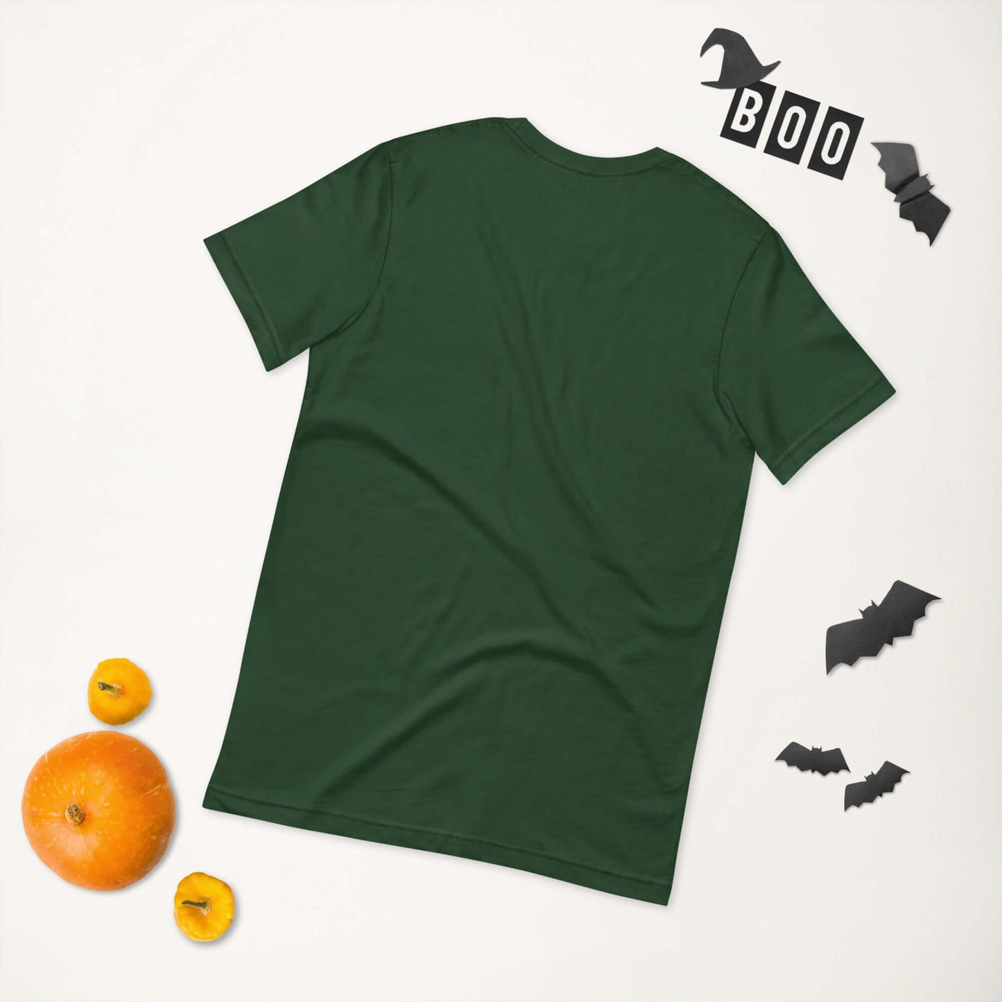 Green unisex t-shirt laid on flat surface, surrounded by Halloween decorations including bats, pumpkins, and a "BOO" sign